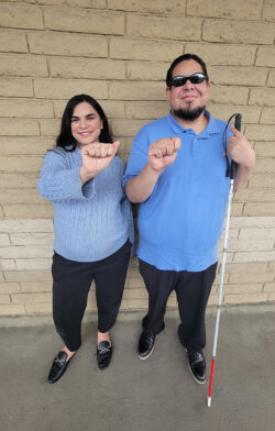 AT Coordinator Team Lizette and Alan signing “A” and “T” in ASL standing outside in front of beige brick wall