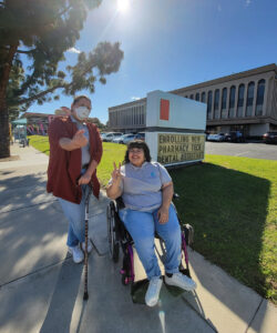 DMC Youth Team Luz and Nelly outside by now enrolling sign on sidewalk with sun shining and trees in background near grass and bulding