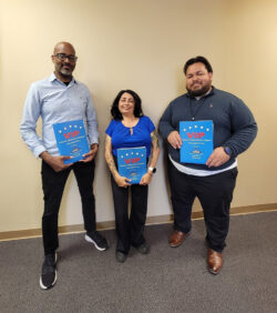 L-R: Andrew Toney, VIP Advisor Los Angeles, Marysol Cuadrado, Lead VIP Advisor and Luis Alvarez, VIP Advisor OC/LA standing together holding VIP information packet standing in front of tan wall inside DMC offices smiling.