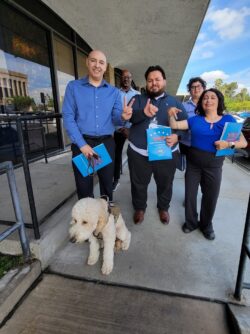 DMC VIP Team on ramp outside building with blue sky and white clouds on sunny day in background. L-R (Front Row) Eric Rodriguez, VIP Advisor, Riverside with “Honey” Service Dog; Luis Alvarez, VIP Advisor OC/LA, Marisol Cuadrado, Lead VIP Advisor – the three of them in the front row signing V-I-P in finger spelling ASL. Back row: Andrew Toney, VIP Advisor Los Angeles and Michelle Ramos, DMC Director of Community Support Services.