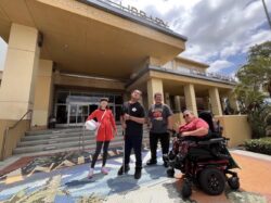 A group of cross-disability youth posing for a photo in front the library. They are smiling and doing poses. In the background, there are signs for “Library” and “City of Anaheim.”
