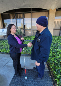Gaby, DMC’s ILS Instructor for the Blind with consumer shaking hands outside as consumer begins journey with DMC for support. Both are smiling as consumer wears blue coat and matching ski cap, Gaby holds mobility cane wearing violet blouse as they stand at base of ramp outside DMC building.