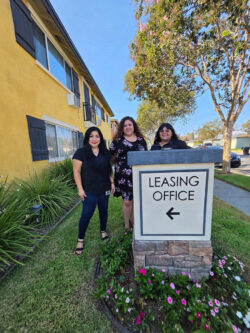 Housing team L-R Monica, Marisol and Paola with leasing sign featuring arrow to the left with pink flowers and shrubs at base, outside of yellow-painted apartment building
