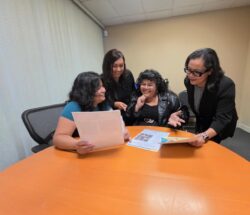 Front office I&R Team together pouring over new flyers and information at table smiling inside DMC office.