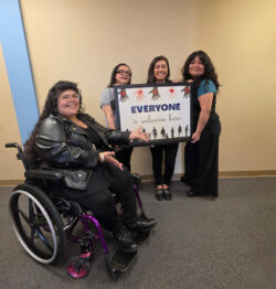 Front office team L-R Nelly, Rosaura, Bianca and Marelli smiling around colorful “EVERYONE IS WELCOME HERE” sign in frame featuring hands and silhouettes of individuals.
