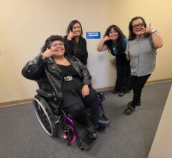 Front office team smiling making a “call me” hand gesture inside DMC offices in front of tan wall with blue reception sign featuring white arrow. L-R: Nelly, Bianca, Mareli and Rosaura.
