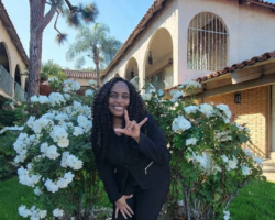 Pamela Torres, Lead Deaf Services Advocate signing ILY in ASL outside with white rosebushes and courtyard buildings behind her.