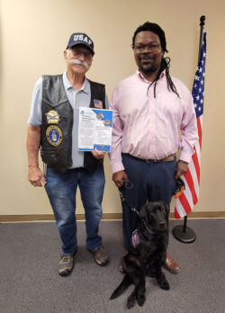 Tony Clement, Veteran and DMC Board President and Griffin Dooley, Veteran and DMC Board Governance with Veterans Independence Program Flyer smiling standing next to U.S. Flag in office with Service dog Vivy.